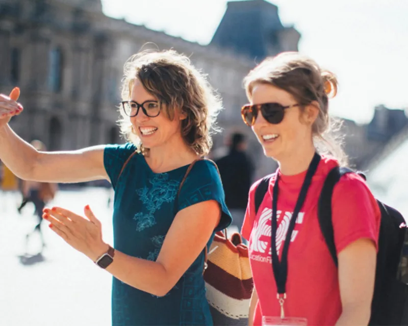 Dos mujeres en una plaza histórica al aire libre, una de ellas con las manos levantadas señalando un monumento al fondo.