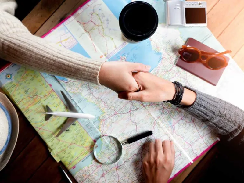 Apretón de manos de dos personas sobre un mapa desplegado en una mesa de madera, con cámara, pasaporte y gafas de sol.