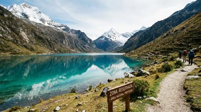 Laguna turquesa en los Andes con letrero de zona reservada y excursionistas caminando por un sendero.