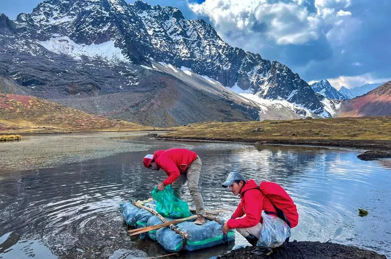 Dos personas con chaquetas rojas recolectan residuos en una laguna de montaña con picos nevados al fondo.